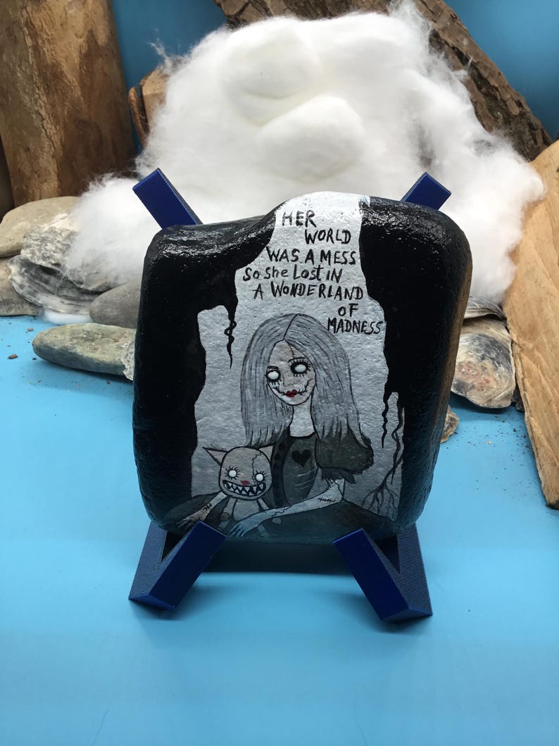 May include: A painted rock featuring a grayscale illustration of a girl and a creature, set against a dark backdrop. The text on the rock reads "Her world was a mess so she lost in a wonderland of madness." The rock is displayed on a blue stand.
