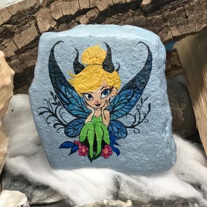 May include: Hand-painted rock art featuring a fairy with blue wings, green dress, and blonde hair. The fairy is sitting on a blue rock with pink flowers.
