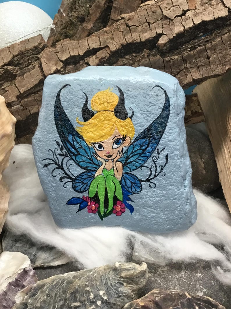 May include: Hand-painted rock art featuring a fairy with blue wings, green dress, and blonde hair. The fairy is sitting on a blue rock with pink flowers.