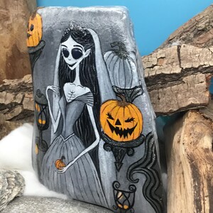 K&ouml;nnte beinhalten: Handbemalte Felskunst mit einer gotischen Braut mit langen schwarzen Haaren, Schleier und Krone. Das Kunstwerk umfasst orangefarbene K&uuml;rbislaternen und dekorative Elemente in Schwarz und Grau, ideal f&uuml;r Halloween-Dekorationen.
