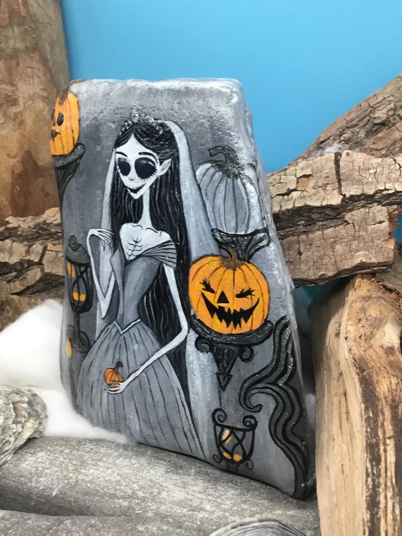 K&ouml;nnte beinhalten: Handbemalte Felskunst mit einer gotischen Braut mit langen schwarzen Haaren, Schleier und Krone. Das Kunstwerk umfasst orangefarbene K&uuml;rbislaternen und dekorative Elemente in Schwarz und Grau, ideal f&uuml;r Halloween-Dekorationen.
