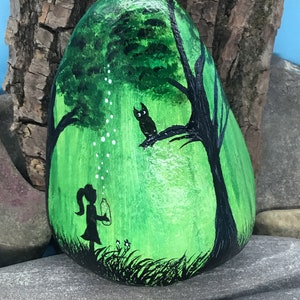 K&ouml;nnte beinhalten: Handbemalter Stein mit einer Darstellung eines M&auml;dchens, das Seifenblasen in einem gr&uuml;nen Wald bl&auml;st, mit einer schwarzen Silhouette einer Eule, die auf einem Ast sitzt.