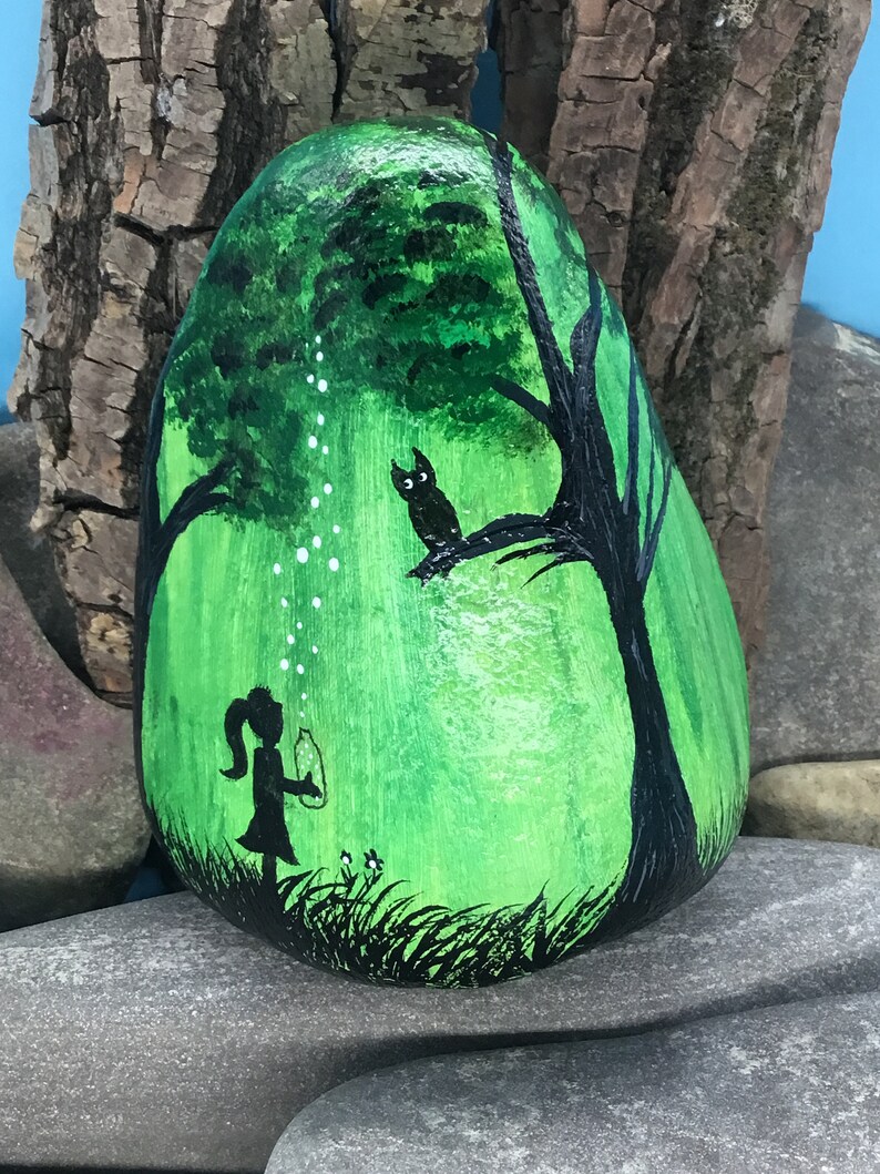 K&ouml;nnte beinhalten: Handbemalter Stein mit einer Darstellung eines M&auml;dchens, das Seifenblasen in einem gr&uuml;nen Wald bl&auml;st, mit einer schwarzen Silhouette einer Eule, die auf einem Ast sitzt.