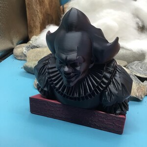 May include: A black bust of a sinister clown with a ruffled collar and a menacing expression. The bust is mounted on a rectangular, maroon base. The clown's hair is styled in a distinctive, pointed shape.