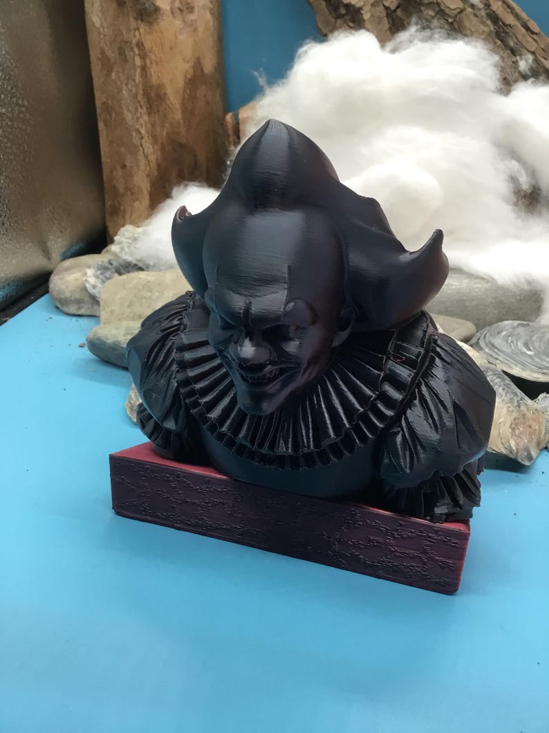 May include: A black bust of a sinister clown with a ruffled collar and a menacing expression. The bust is mounted on a rectangular, maroon base. The clown's hair is styled in a distinctive, pointed shape.
