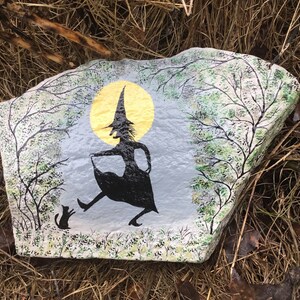 May include: A painted rock with a silhouette of a witch in a pointed hat and a black cat. The witch is walking with a basket in her hand and a full moon is behind her. The rock is painted with green and white trees and a gray background.