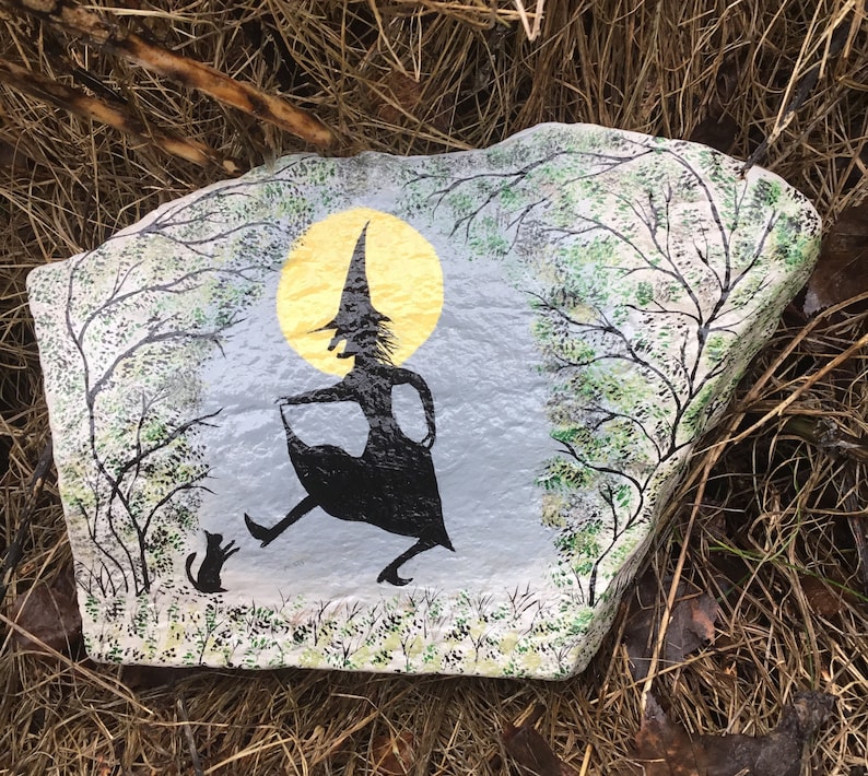 May include: A painted rock with a silhouette of a witch in a pointed hat and a black cat. The witch is walking with a basket in her hand and a full moon is behind her. The rock is painted with green and white trees and a gray background.
