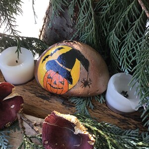May include: A painted rock featuring a witch's hat and a jack-o'-lantern face. The rock is surrounded by greenery and two white candles.