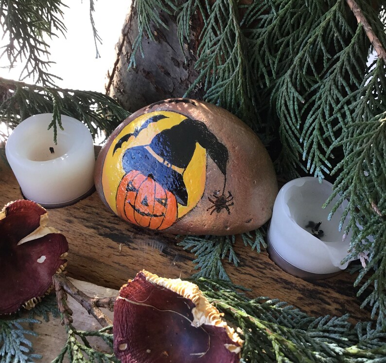 May include: A painted rock featuring a witch's hat and a jack-o'-lantern face. The rock is surrounded by greenery and two white candles.
