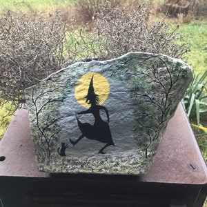 May include: A painted rock depicting a witch in a black dress and hat, with a black cat, standing in front of a yellow moon and trees.