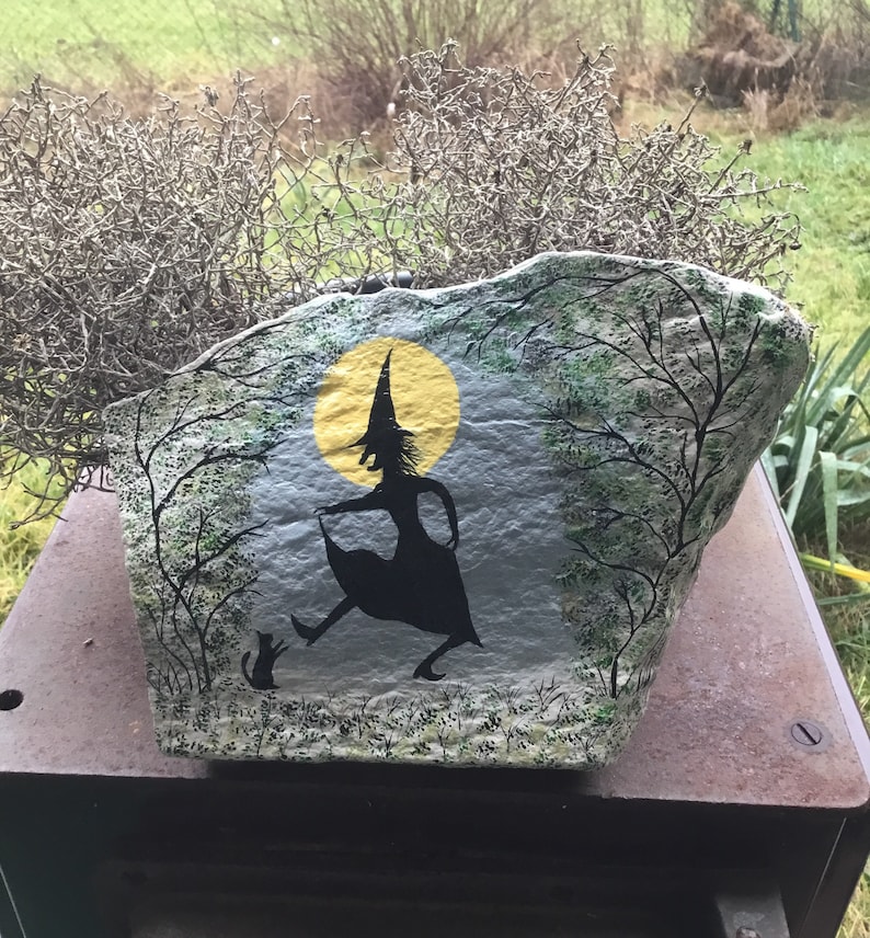 May include: A painted rock depicting a witch in a black dress and hat, with a black cat, standing in front of a yellow moon and trees.