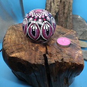Op de afbeelding: Een zwarte bol met roze en witte stippen en swirls erop geschilderd. De bol ligt op een boomstronk met een kleine roze kaars op de stronk.