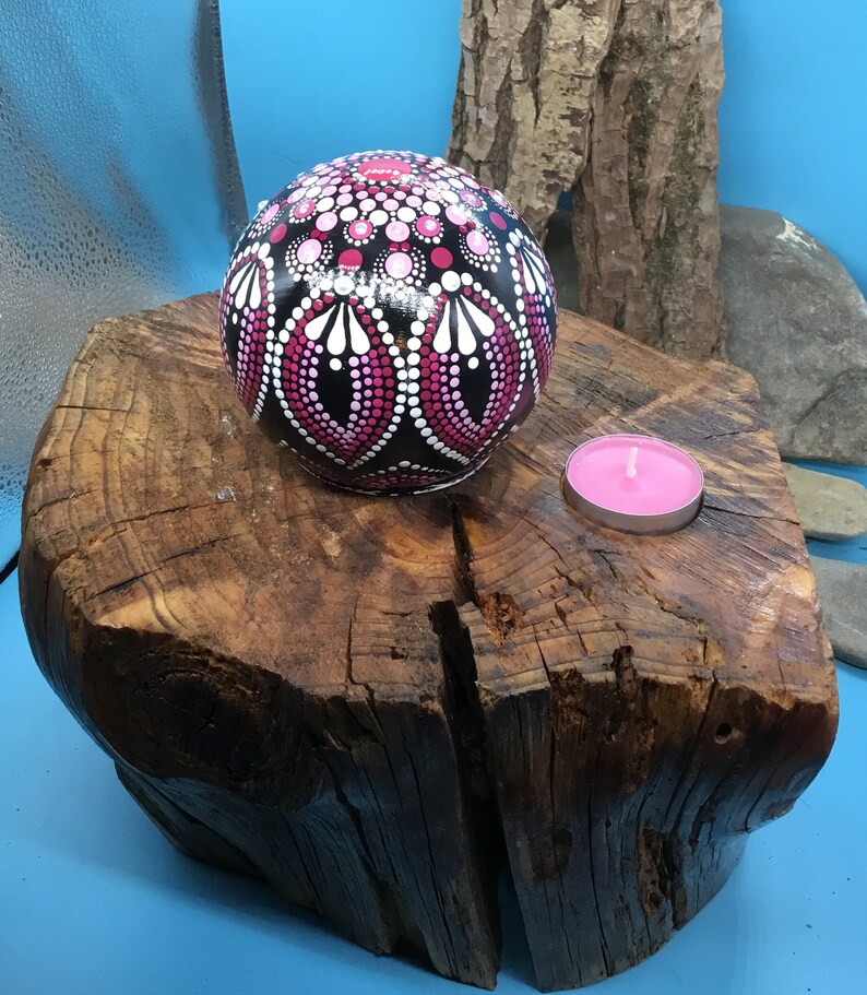 Op de afbeelding: Een zwarte bol met roze en witte stippen en swirls erop geschilderd. De bol ligt op een boomstronk met een kleine roze kaars op de stronk.