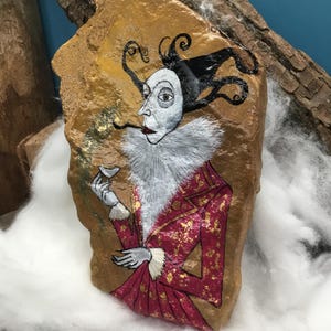 Op de afbeelding: Handgeschilderd kunstwerk op een steen dat een figuur afbeeldt in een rood en goudkleurig jack, witte kraag en zwarte hoed met krullende accenten. De figuur houdt een klein glas vast. De steen heeft een gouden achtergrond.