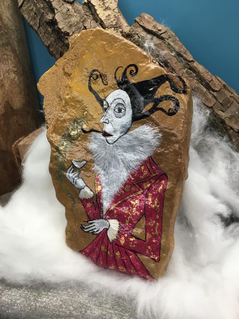 Op de afbeelding: Handgeschilderd kunstwerk op een steen dat een figuur afbeeldt in een rood en goudkleurig jack, witte kraag en zwarte hoed met krullende accenten. De figuur houdt een klein glas vast. De steen heeft een gouden achtergrond.