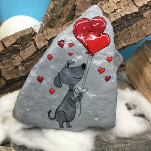 May include: A hand-painted gray rock with a cartoon dog holding two red heart-shaped balloons. The dog is surrounded by smaller red hearts.