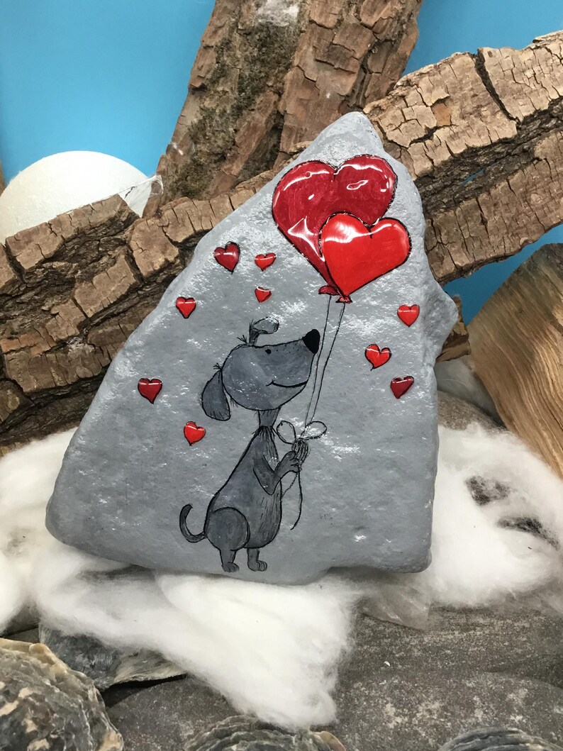 May include: A hand-painted gray rock with a cartoon dog holding two red heart-shaped balloons. The dog is surrounded by smaller red hearts.