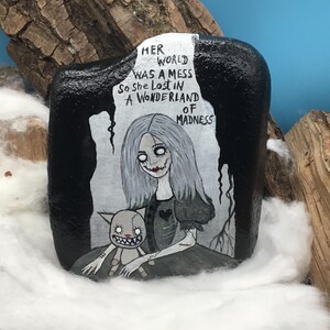 May include: Hand-painted rock art featuring a gothic Alice in Wonderland theme. The artwork depicts a pale-skinned girl with gray hair and a doll, with the text "Her world was a mess so she lost in a wonderland of madness."