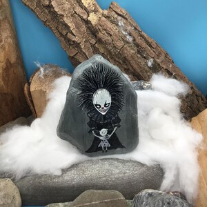 May include: A grey painted rock featuring a detailed illustration of a pale, wide-eyed clown with spiky black hair and a ruffled collar, holding a small white doll with black polka dots. The clown has a sharp, toothy grin and red nose accents.
