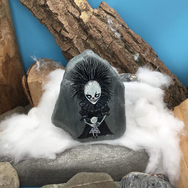 May include: A grey painted rock featuring a detailed illustration of a pale, wide-eyed clown with spiky black hair and a ruffled collar, holding a small white doll with black polka dots. The clown has a sharp, toothy grin and red nose accents.