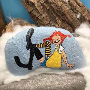 May include: A light blue painted rock featuring a hand-painted illustration of a character with red hair in pigtails, wearing a yellow dress, blue polka-dot shorts, yellow and black striped stockings, and black boots, sitting in a relaxed pose.