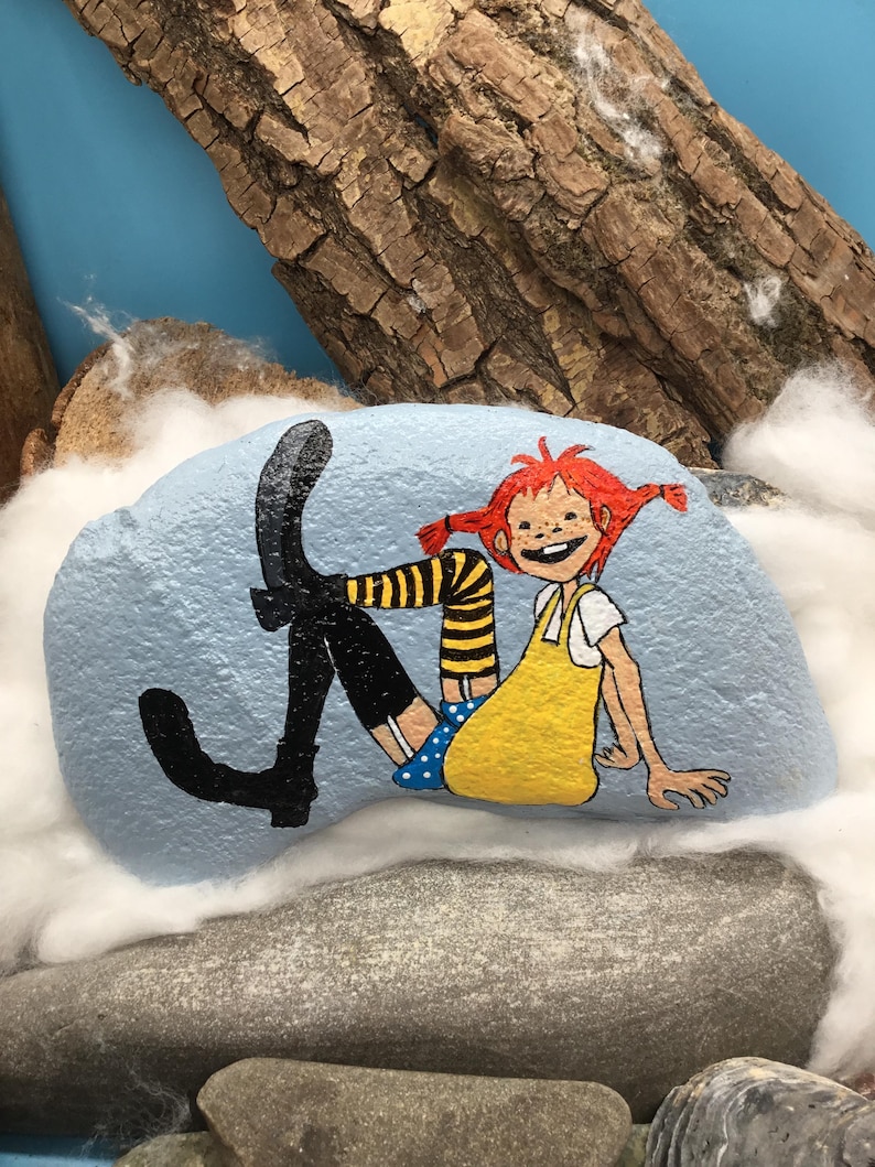 May include: A light blue painted rock featuring a hand-painted illustration of a character with red hair in pigtails, wearing a yellow dress, blue polka-dot shorts, yellow and black striped stockings, and black boots, sitting in a relaxed pose.
