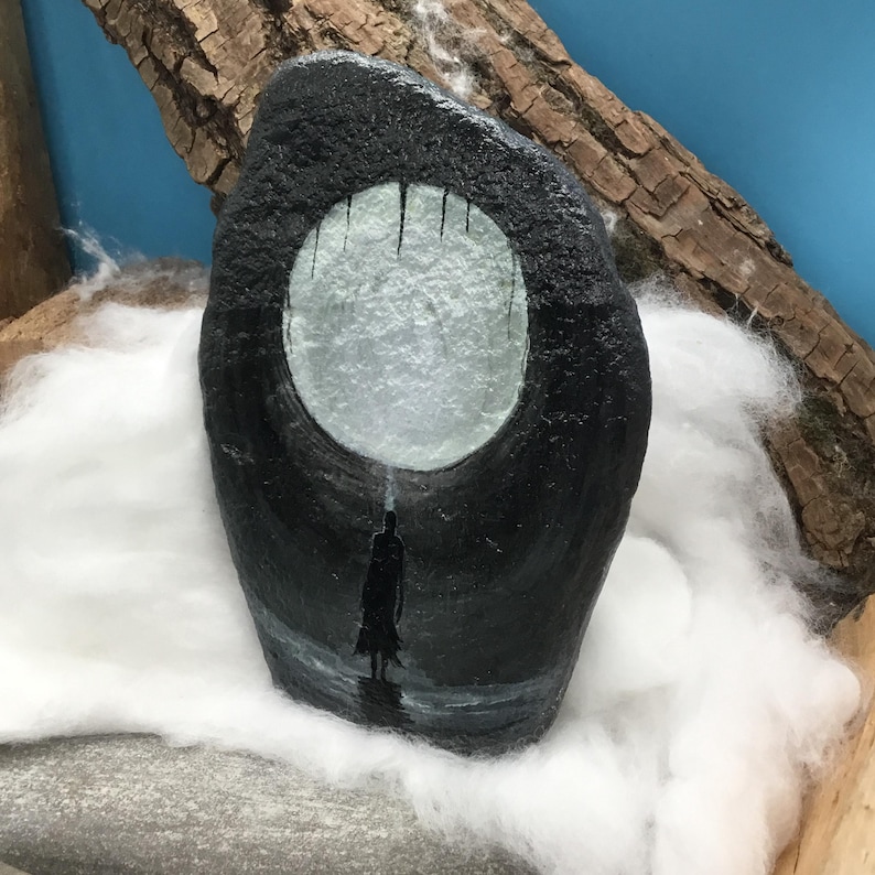 Pu&ograve; includere: Una pietra nera dipinta a mano con un'apertura circolare che raffigura una figura silhouette in piedi nell'acqua sotto un cielo illuminato dalla luna. L'opera d'arte &egrave; ambientata su uno sfondo di cotone bianco e un pezzo di legno.