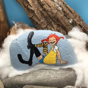 May include: A light blue painted rock featuring a hand-painted illustration of a character with bright red hair in pigtails, wearing a yellow dress, blue polka-dot shorts, and black boots with yellow and black striped stockings.