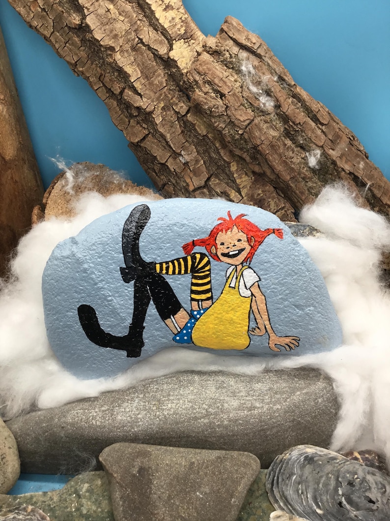 May include: A light blue painted rock featuring a hand-painted illustration of a character with bright red hair in pigtails, wearing a yellow dress, blue polka-dot shorts, and black boots with yellow and black striped stockings.