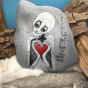 May include: A gray painted rock with a skeleton holding a red heart. The skeleton has large eyes and a stitched mouth. Text on the rock reads "I do find most people to be awfully boring."