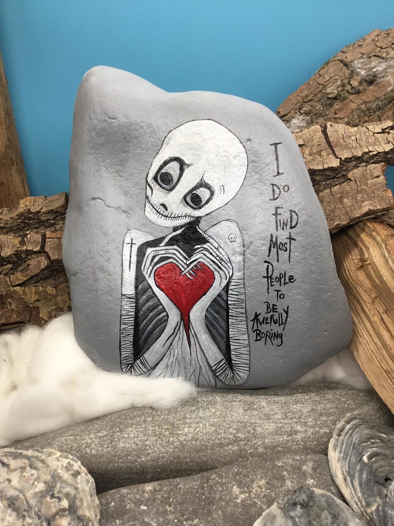 May include: A gray painted rock with a skeleton holding a red heart. The skeleton has large eyes and a stitched mouth. Text on the rock reads "I do find most people to be awfully boring."