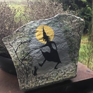 May include: Hand-painted rock art depicting a witch in silhouette flying with a cat under a yellow moon. The background is a gray and green forest scene.