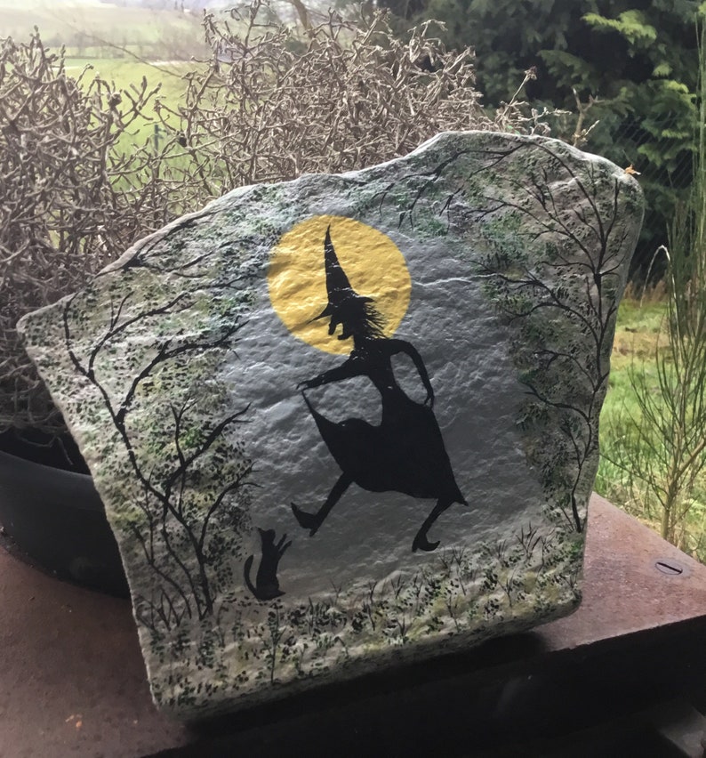 May include: Hand-painted rock art depicting a witch in silhouette flying with a cat under a yellow moon. The background is a gray and green forest scene.
