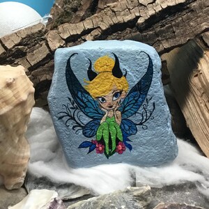 May include: Hand-painted rock art featuring a fairy with blue wings, green dress, and blonde hair. The fairy is sitting on a blue background with pink flowers.