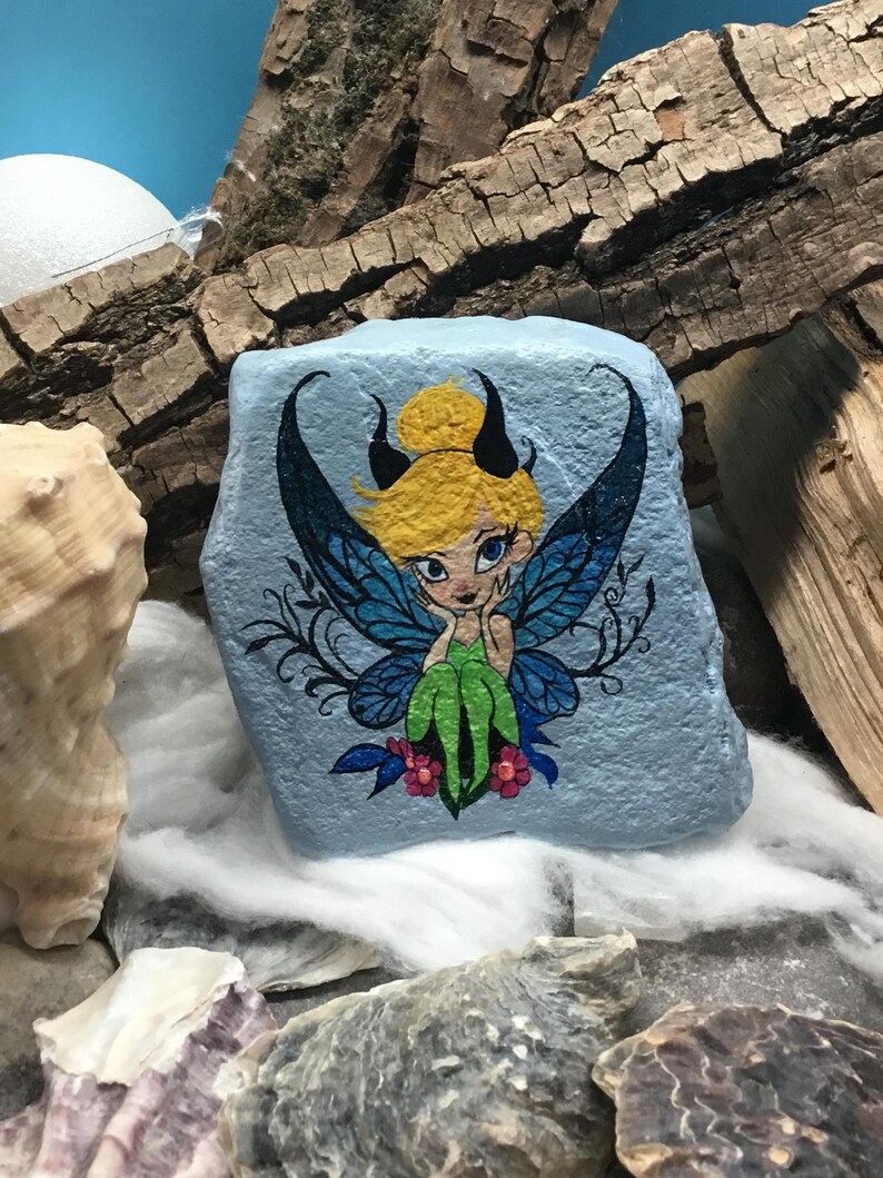 May include: Hand-painted rock art featuring a fairy with blue wings, green dress, and blonde hair. The fairy is sitting on a blue background with pink flowers.