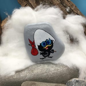 K&ouml;nnte beinhalten: Ein handbemalter, grauer Stein zeigt einen Cartoon-K&uuml;ken-Charakter mit einem wei&szlig;en Eierschalenhut, blauen Augen und einem roten Sack. Das K&uuml;ken ist schwarz mit einem gelben Schnurrbart und tr&auml;gt einen kleinen Stock. Der Stein liegt auf einem Bett aus wei&szlig;em Flaum.