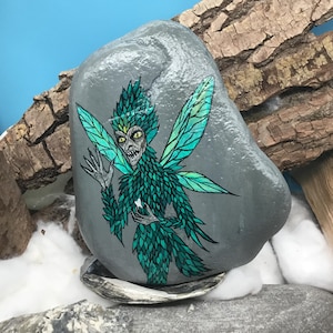 May include: A hand-painted gray rock features a detailed illustration of a fairy. The fairy has green and teal wings and matching leafy attire. The fairy's face is pale with yellow eyes and a menacing expression. The rock rests on a seashell.