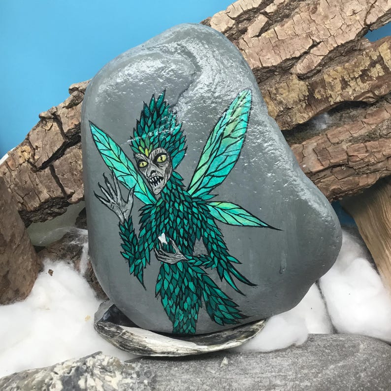 May include: A hand-painted gray rock features a detailed illustration of a fairy. The fairy has green and teal wings and matching leafy attire. The fairy's face is pale with yellow eyes and a menacing expression. The rock rests on a seashell.