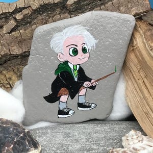May include: Hand-painted rock art featuring a cartoon character with white hair, green eyes, and a green tie, holding a wand. The character wears a black robe and is depicted on a gray rock. The art is set against a backdrop of wood and seashells.