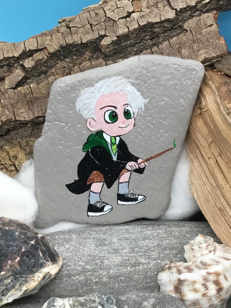 May include: Hand-painted rock art featuring a cartoon character with white hair, green eyes, and a green tie, holding a wand. The character wears a black robe and is depicted on a gray rock. The art is set against a backdrop of wood and seashells.