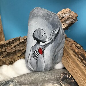 K&ouml;nnte beinhalten: Ein handbemalter grauer Stein zeigt eine Alienfigur mit einem roten Herzen und freiliegendem Brustkorb. Das Kunstwerk ist in Schwarz, Wei&szlig; und Rot gehalten, wobei der Au&szlig;erirdische die Hand ausstreckt. Der Stein wird mit Treibholz und anderen Steinen ausgestellt.