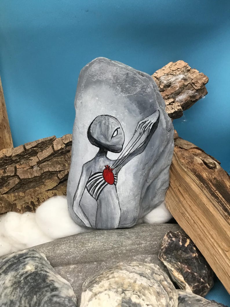 K&ouml;nnte beinhalten: Ein handbemalter grauer Stein zeigt eine Alienfigur mit einem roten Herzen und freiliegendem Brustkorb. Das Kunstwerk ist in Schwarz, Wei&szlig; und Rot gehalten, wobei der Au&szlig;erirdische die Hand ausstreckt. Der Stein wird mit Treibholz und anderen Steinen ausgestellt.