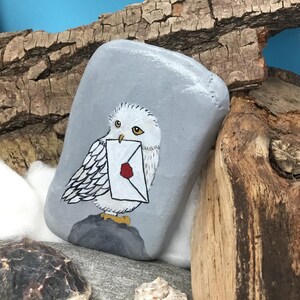 May include: A hand-painted, grey stone features a white owl carrying a letter with a red wax seal. The owl is perched on a dark grey base. The background includes wood, cotton, and seashells, with a blue sky backdrop.