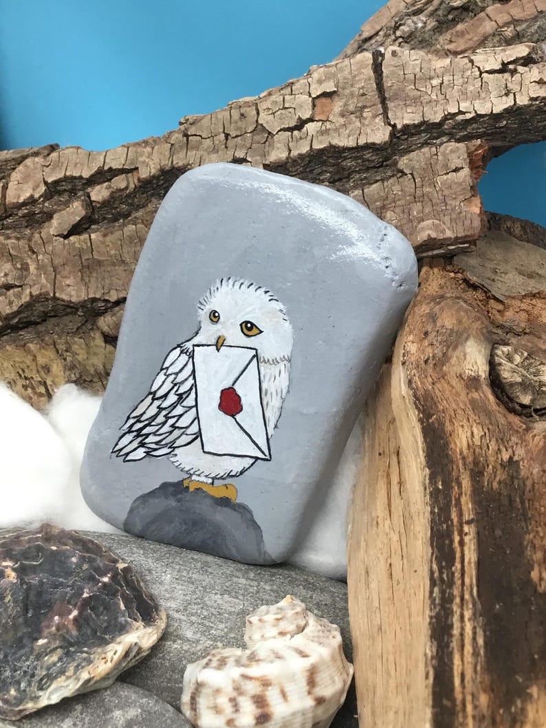 May include: A hand-painted, grey stone features a white owl carrying a letter with a red wax seal. The owl is perched on a dark grey base. The background includes wood, cotton, and seashells, with a blue sky backdrop.