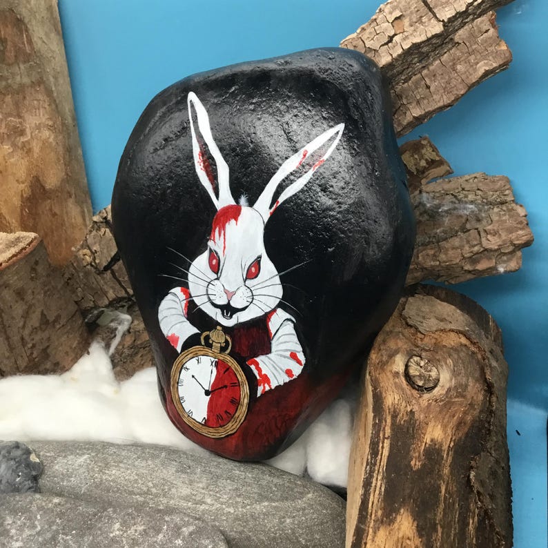 May include: A hand-painted black rock depicts a white rabbit with red eyes and blood-splattered bandages. The rabbit holds a gold pocket watch with a red and white face. The artwork is inspired by the White Rabbit from Alice in Wonderland.