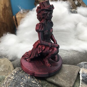 May include: A 3D printed figurine of a person kneeling on a circular base, wearing a top hat with goggles, a corset, and a long skirt. The figure is finished in a metallic dark red and black color scheme, showing intricate textures and details.