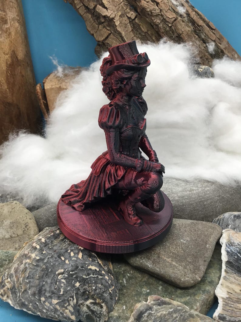 May include: A 3D printed figurine of a person kneeling on a circular base, wearing a top hat with goggles, a corset, and a long skirt. The figure is finished in a metallic dark red and black color scheme, showing intricate textures and details.