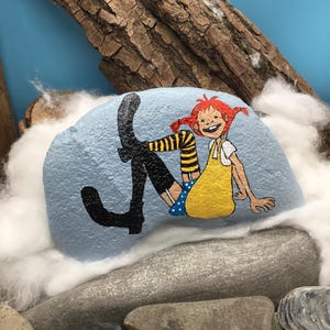 May include: A light blue painted rock featuring a hand-painted illustration of a character with red hair in pigtails, wearing a yellow dress, blue polka dot shorts, and black boots with yellow and black striped stockings, sitting in a relaxed pose.