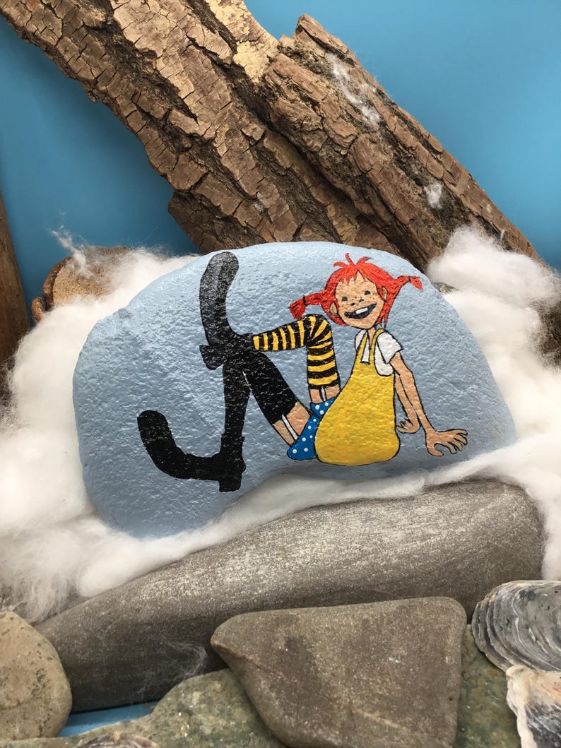 May include: A light blue painted rock featuring a hand-painted illustration of a character with red hair in pigtails, wearing a yellow dress, blue polka dot shorts, and black boots with yellow and black striped stockings, sitting in a relaxed pose.