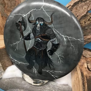 May include: A circular, grey painted wooden plaque depicting a Minotaur warrior with an axe, set against a backdrop of lightning. The Minotaur has blue eyes and black fur. The plaque is displayed on a stone surface.